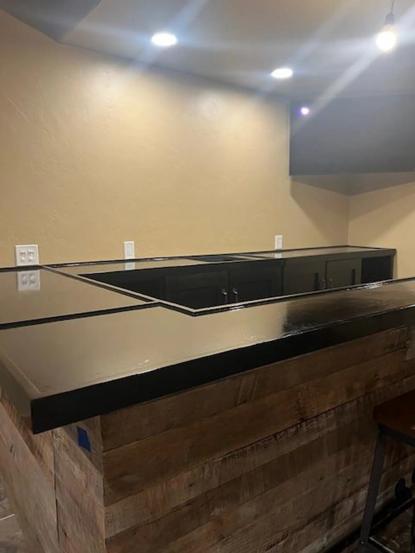 Angled view of custom black-painted wood bar top with barnwood face, showing full L-edge wrap detail and overhead lighting.