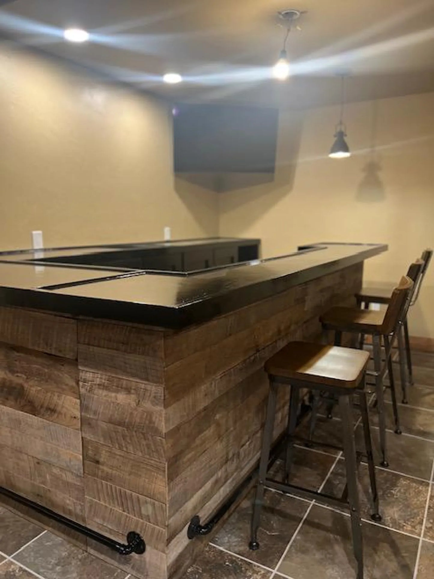 Close-up of U-shaped bar corner showing reclaimed wood texture and black foot rail against tile flooring.