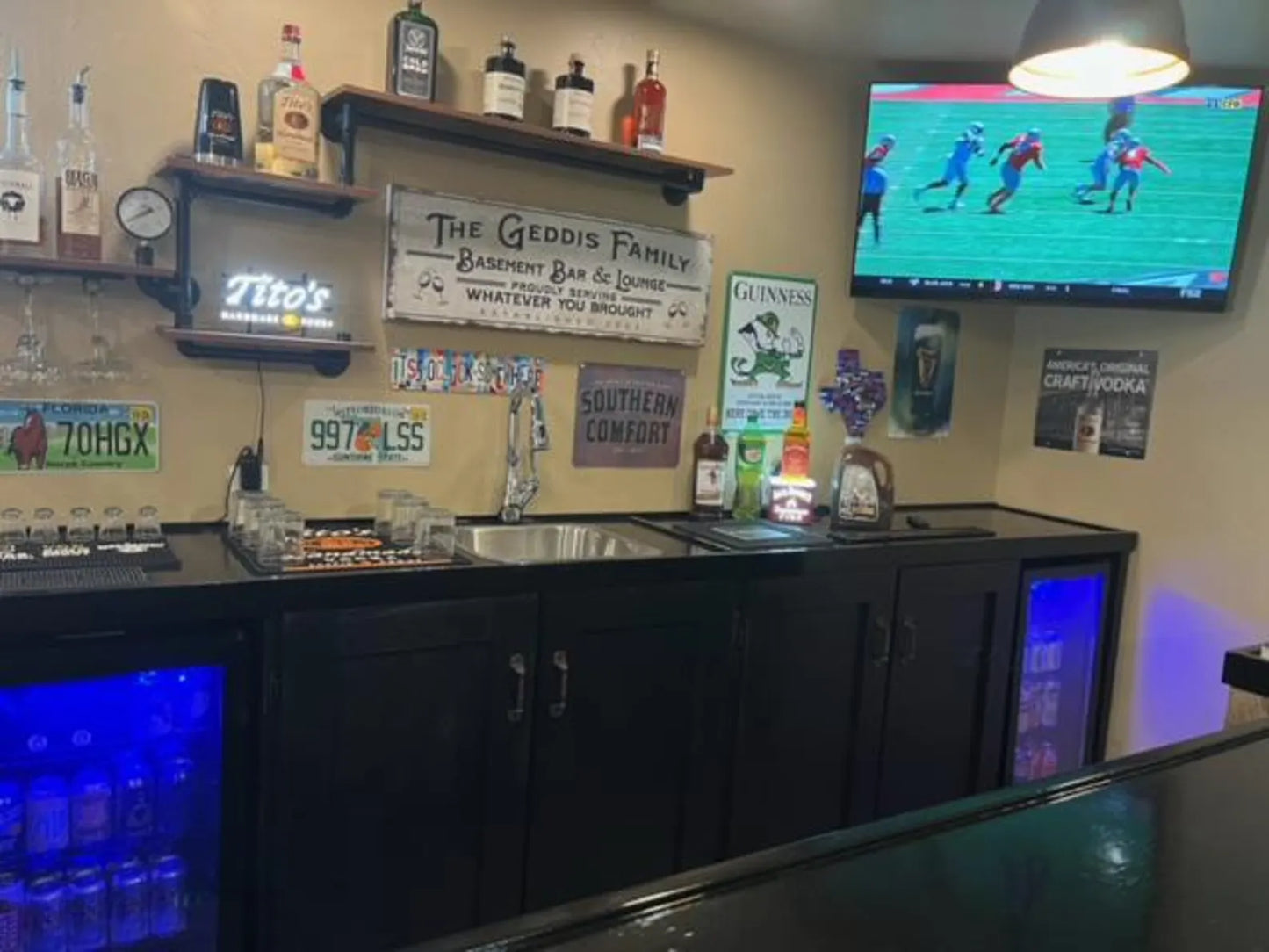 Complete basement bar setup with black cabinets, dual glass-door fridges, overhead shelving, and wall-mounted TV.