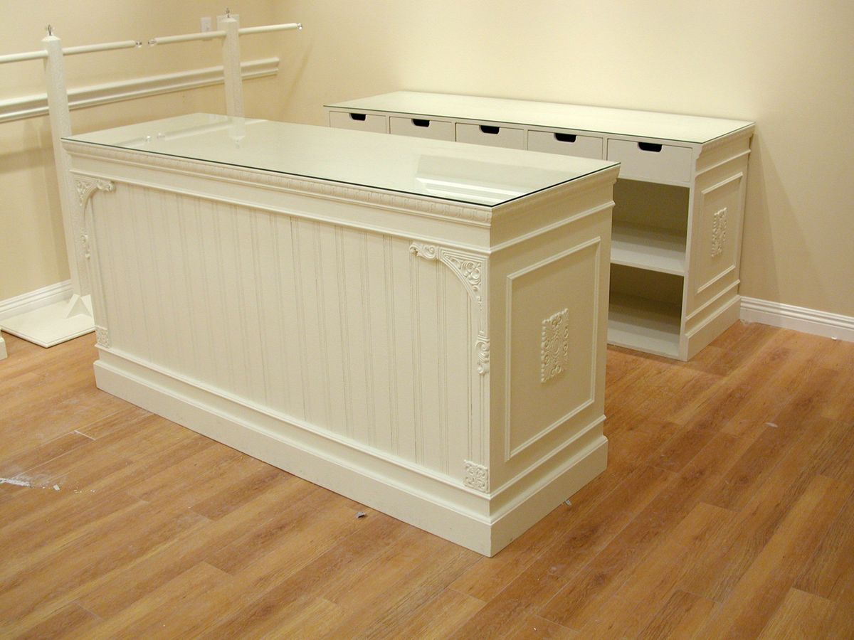 An off-white wooden display case or retail counter with a glass top, shown in a room with a wood floor and beige walls, with another matching cabinet in the background.