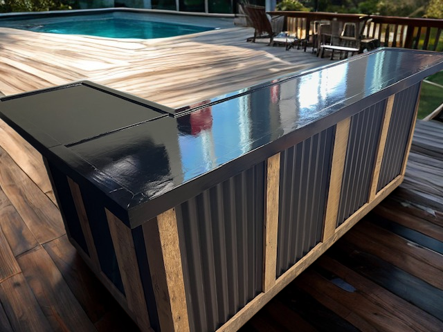 L shaped metal and wood bar by Red Cloak Wood Designs on a wooden deck near a pool, featuring rustic reclaimed wood frame and black corrugated metal panels.