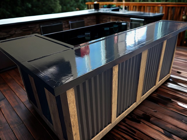 L shaped metal and reclaimed wood bar by Red Cloak Wood Designs set on a wet deck with built-in seating and outdoor kitchen in the background.