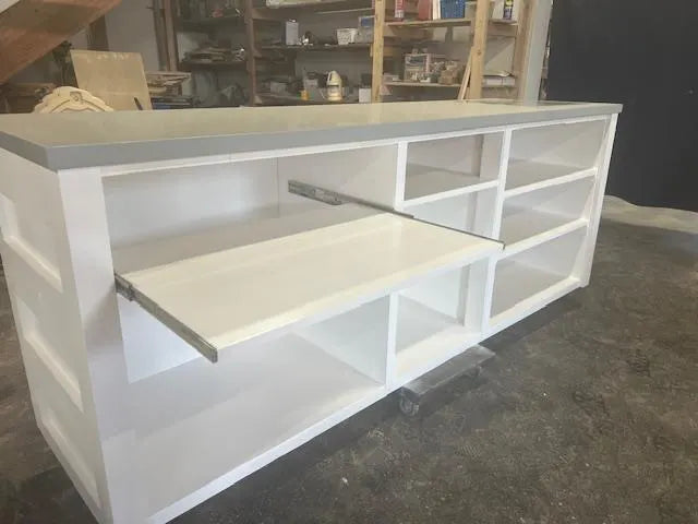 the back of a white custom wood checkout counter with a gray countertop, shown in a workshop or warehouse setting with shelves and tools in the background.