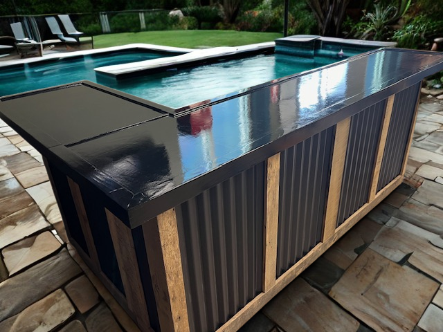 Rustic L shaped metal and wood bar by Red Cloak Wood Designs placed on stone patio beside a backyard pool, built with corrugated metal panels and reclaimed wood trim.
