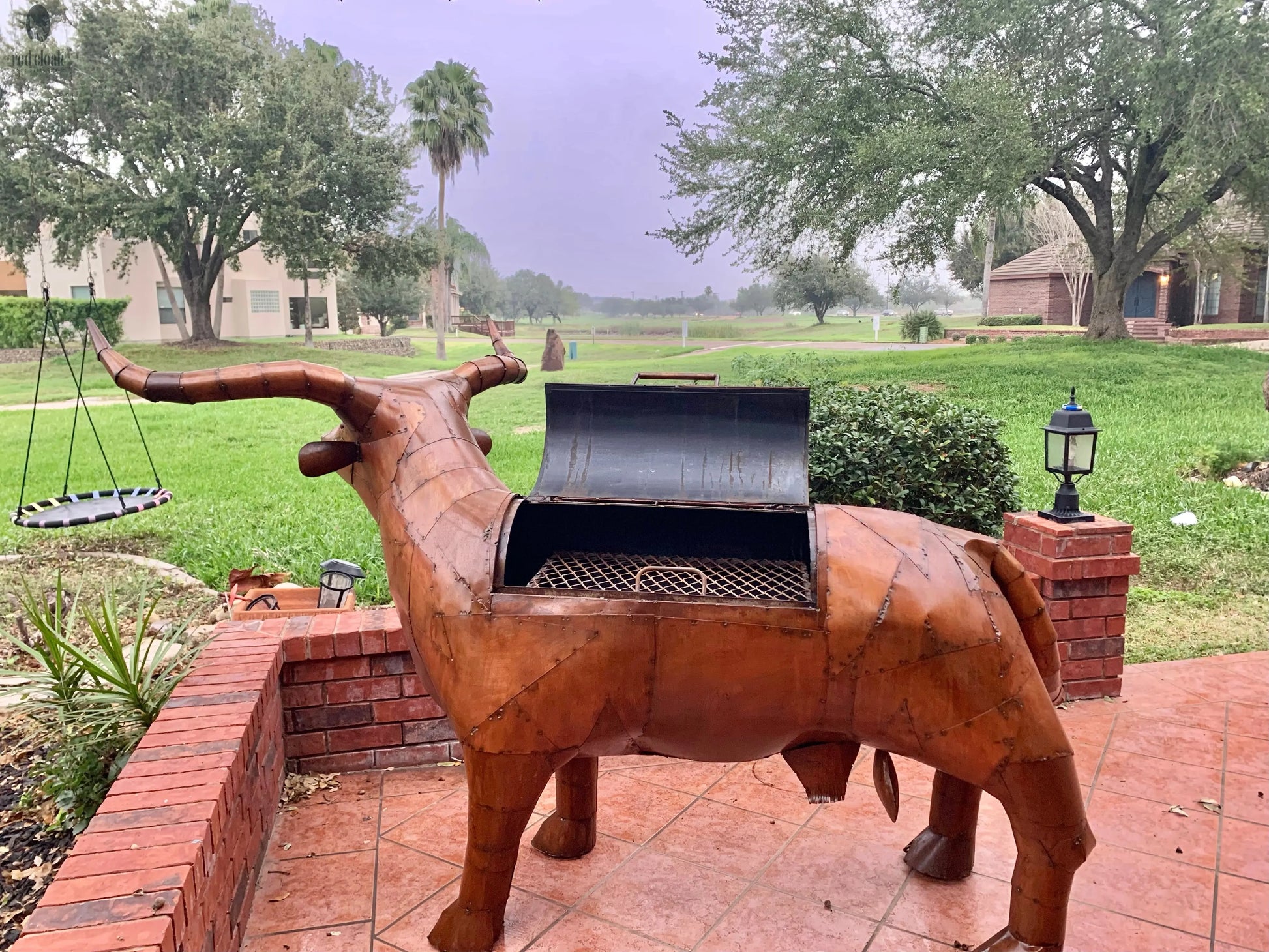 full view of copper longhorn BBQ grill with lid open, set on red brick patio in backyard with greenery. Handmade by Red Cloak Wood Designs