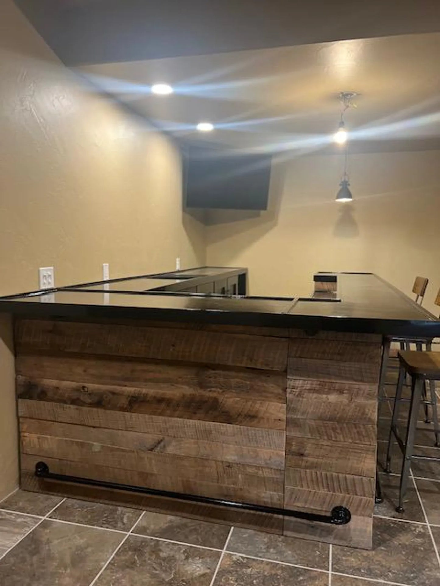 Reclaimed barnwood front panel of U-shaped indoor bar with overhang and black foot rail, built into basement barroom with tile floors.
