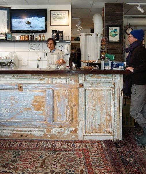 A rustic, distressed wooden checkout counter in a cafe setting, with a woman working behind it and a man standing to the right. The counter features peeling blue and beige paint, set against a tiled wall with a TV and coffee equipment.