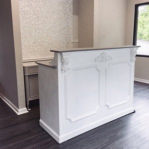 A white checkout counter with decorative paneling and a gray countertop, shown in a room with dark wood floors and beige walls.
