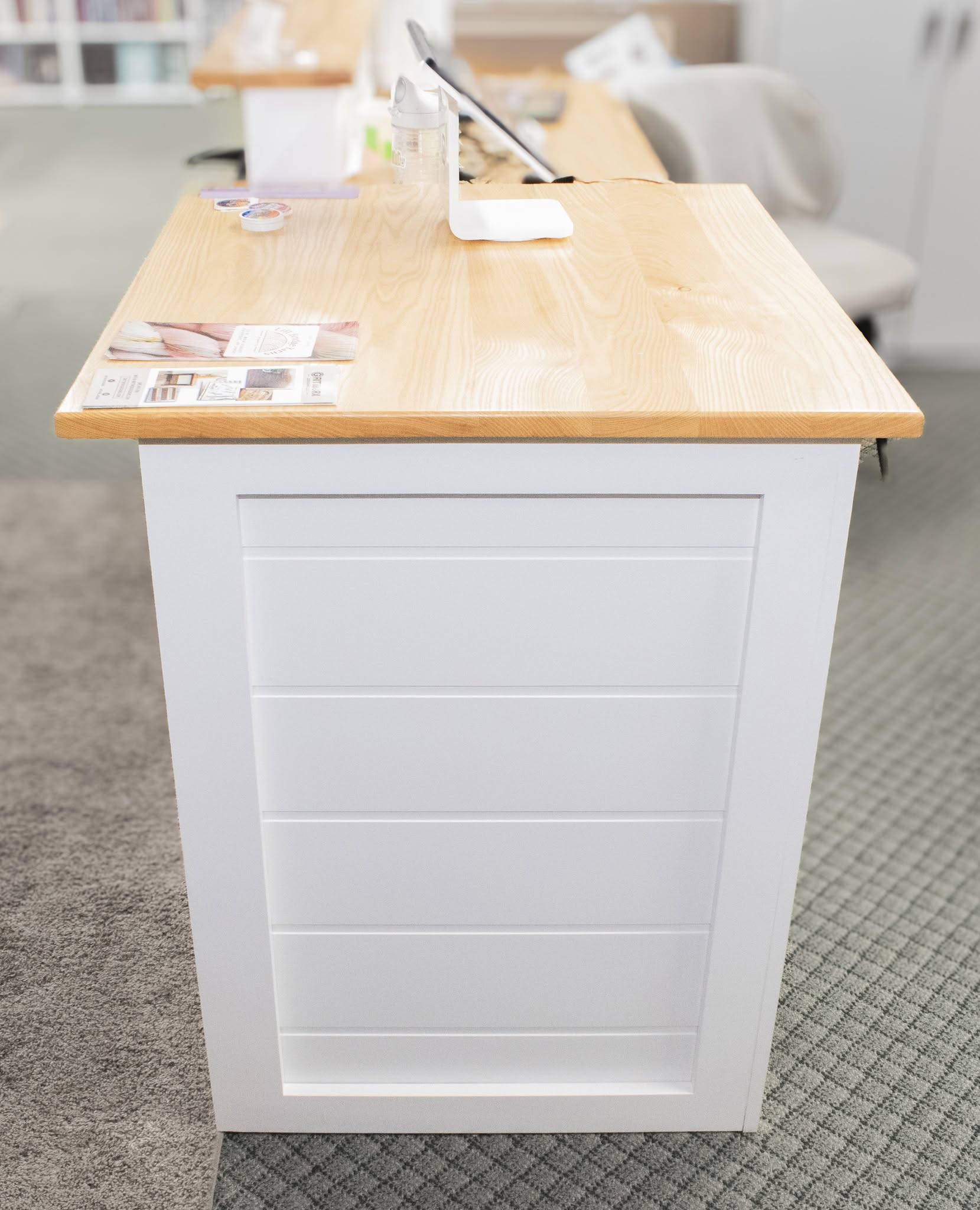 A white reception desk with a light natural wood top, featuring a paneled front and a small tabletop organizer on top.