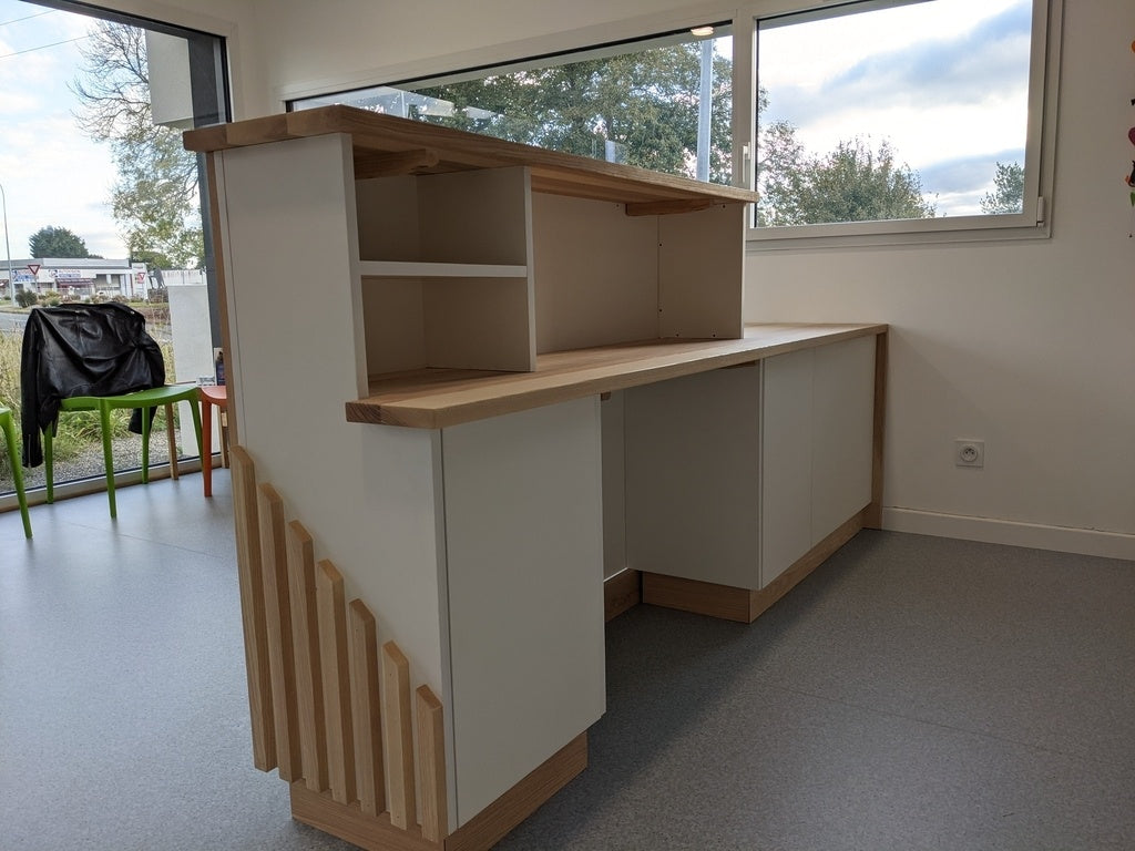 A modern white and light wood reception desk with open shelving and a slatted decorative panel on the left side, set in a room with gray flooring and large windows.