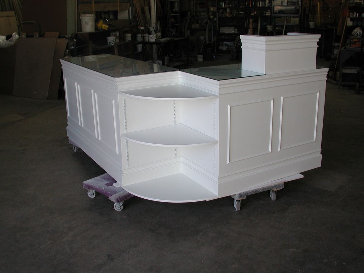 A white corner reception desk or checkout counter with built-in shelving, a glass top, and caster wheels, photographed in a workshop or warehouse setting.