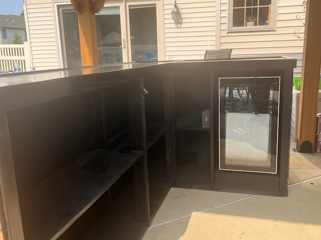 Dark brown outdoor L-shaped bar counter with a refrigerator and open shelving, shown on a concrete patio in front of a house.