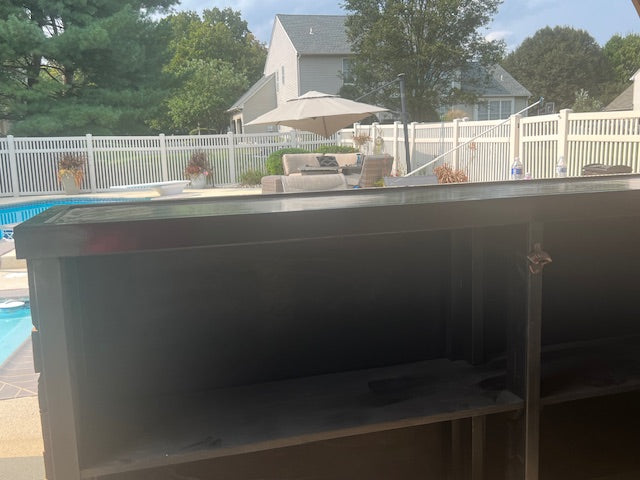 A custom wood outdoor bar with open shelving, photographed in a backyard setting with a pool, white fence, patio umbrella, and house visible in the background.
