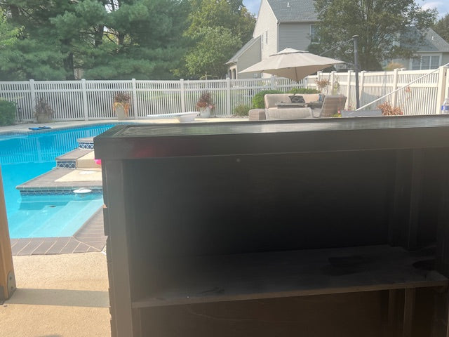 A custom made outdoor wooden bar  in the foreground, photographed in a backyard setting with a swimming pool, white fence, trees, and patio furniture visible in the background.