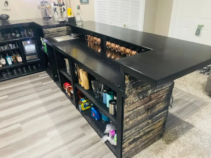 Interior view of L shaped bar with built-in bartender station, black bar top, and rustic shelving for glassware and mixers by Red Cloak