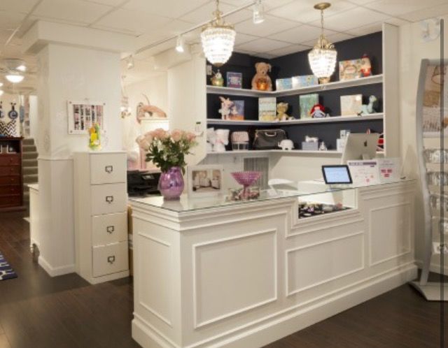 A white wooden retail counter with a glass top, set in a boutique interior with dark wood floors, shelving, and hanging pendant lights.