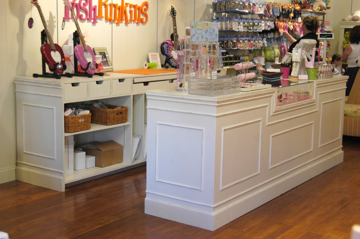 A white wooden retail display counter and matching storage cabinet in a boutique setting, with guitars on display, merchandise racks, and a wood floor.