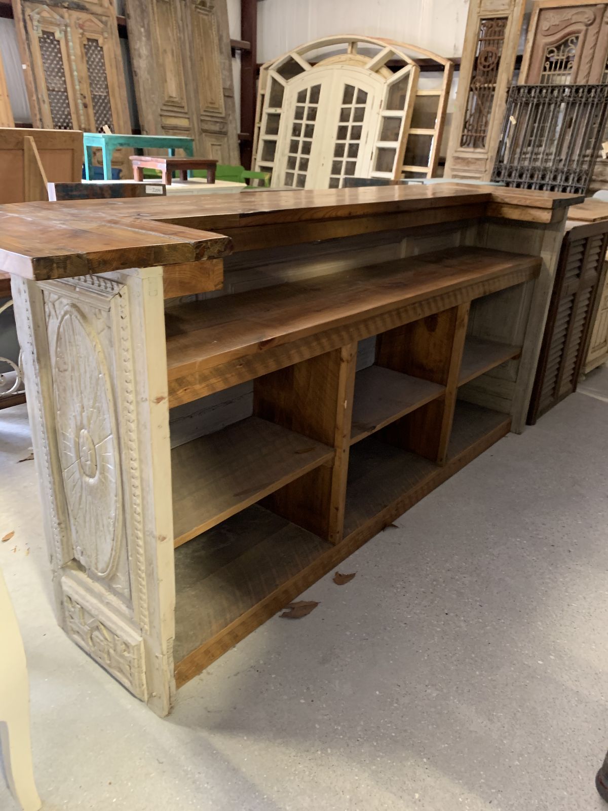 A rustic wooden counter or bar with carved decorative side panels and open shelving, photographed in a furniture store or workshop setting.