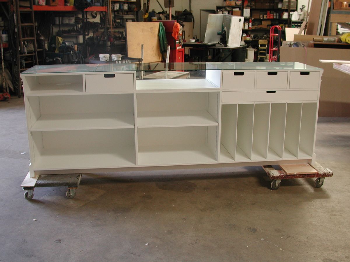 A white wooden counter or display case with a glass top, multiple open shelves, drawers, and storage cubbies, shown on a wheeled dolly in a workshop or warehouse setting.