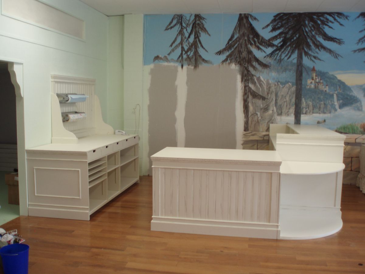 A white curved reception desk, checkout counter, or retail counter in a showroom with a wood floor, a matching white storage cabinet on the left, and a large scenic mural on the back wall.