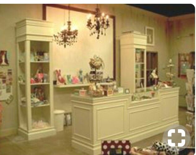 A vintage-style boutique interior with cream-colored display cabinets, a central counter, and hanging chandeliers, displaying various small items and dolls.