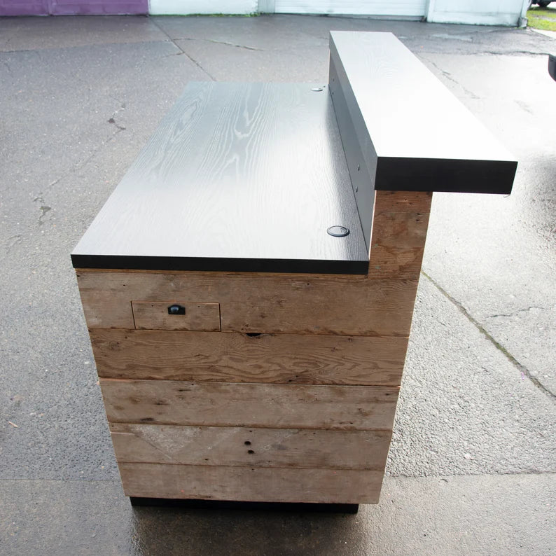 Angled side view of a reclaimed wood reception desk with cord grommets, storage drawer, and black worktop. Handmade by Red Cloak Home