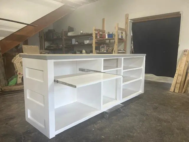 the back of a white handmade wood retail counter with multiple shelves and a pull-out tray, shown in a workshop or garage setting.