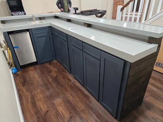 L-shaped custom wood bar with dark blue-gray shaker-style cabinets, a white countertop, and a wood-look side panel, installed in a home bar setting with wood flooring and an ice maker visible.