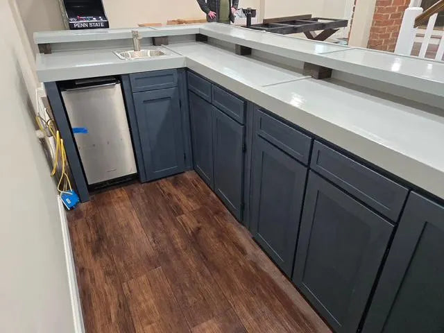 A L-shaped custom wood bar with dark slate blue cabinets, a gray bartop, a built-in stainless steel mini refrigerator, and brown wood-look flooring.