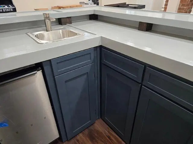 L-shaped home bar cabinets with dark slate blue shaker-style doors and a gray bartop, featuring a built-in stainless steel sink and a stainless steel ice maker on the left.
