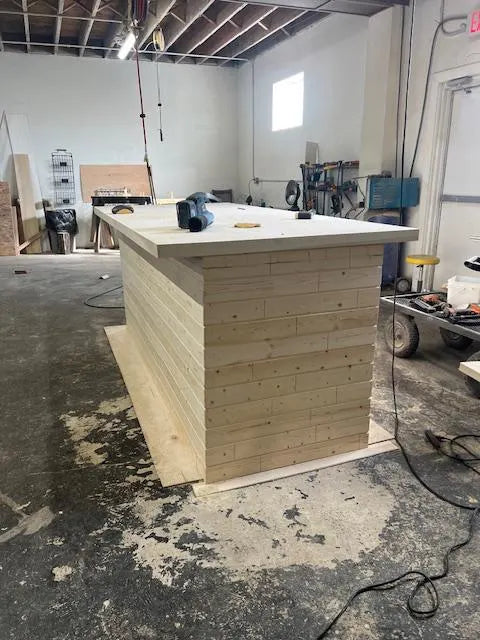 A custom wooden bar with a light natural wood plank base and a white countertop, shown in a workshop or construction site.