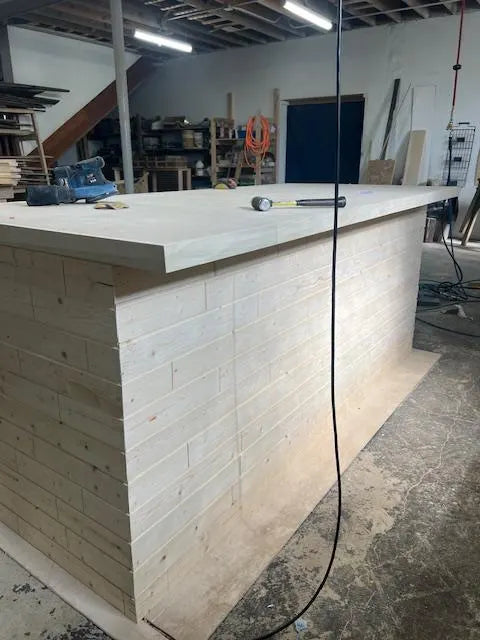 A custom light wood home bar with a thick solid top, shown in a workshop setting with tools on top and a hanging power cord in the foreground.