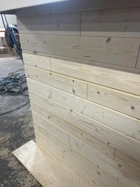 Light natural wood paneling installed on a wall or furniture side, photographed in a workshop or construction setting with concrete floor and cables visible in the background.