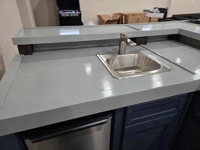 A gray bartop with an integrated stainless steel sink and faucet, installed above dark blue bar cabinets in custom home wood bar.