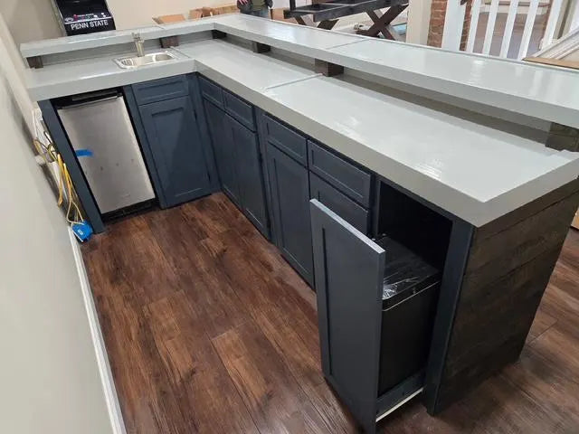 L-shaped custom home wood bar with dark slate blue cabinets, a gray bartop, and a dark wood-look front panel, installed in a home bar with wood-look flooring.