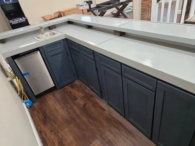 L-shaped custom wood bar cabinetry with dark gray shaker-style lower cabinets and a glossy gray bartop, shown in a room with wood flooring and a built-in stainless steel ice maker.