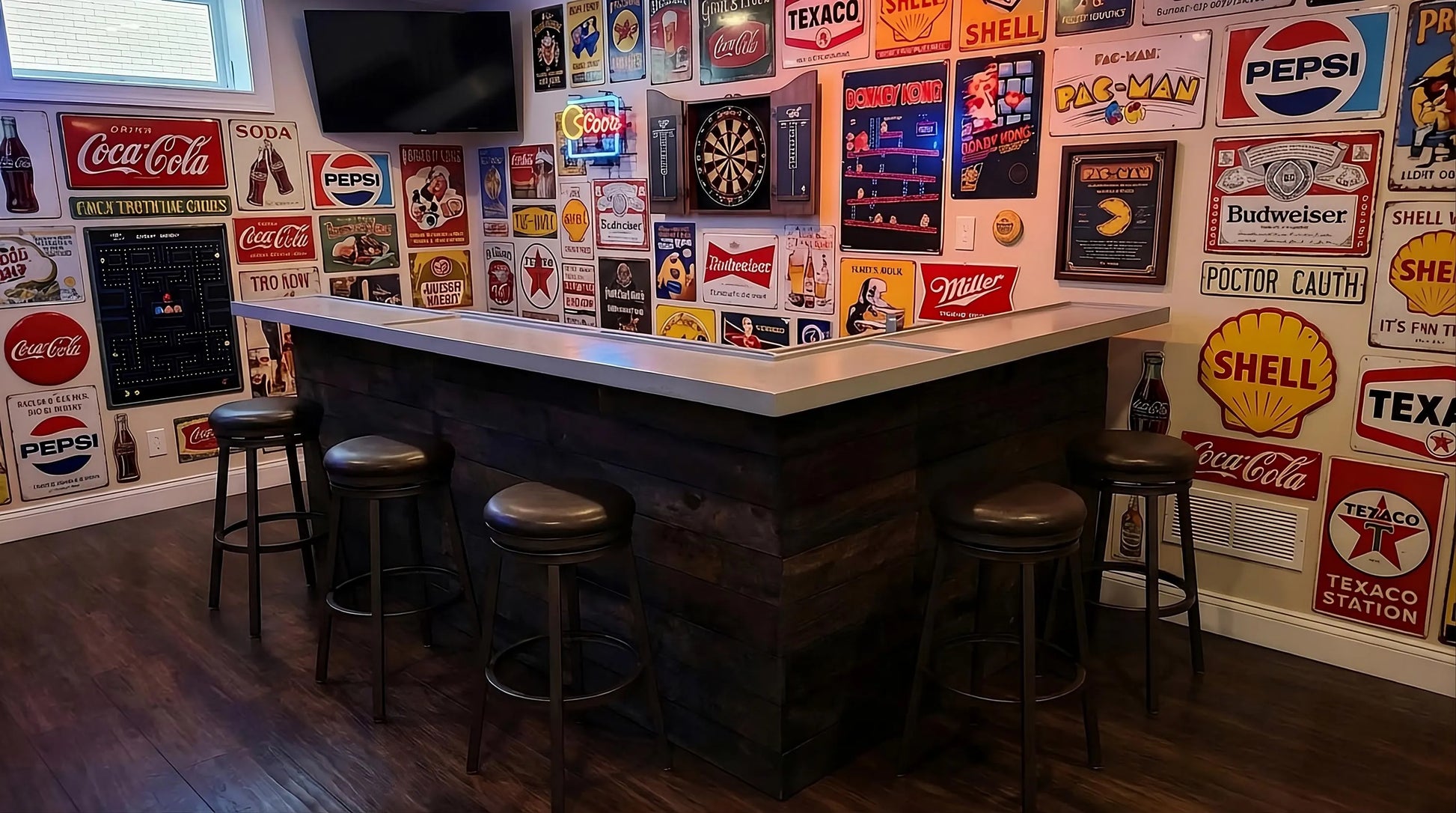 A rustic wood bar with a gray bartop and dark wood base, surrounded by six bar stools in a room decorated with numerous vintage-style Coca-Cola, Pepsi, Texaco, and Shell signs on the walls.