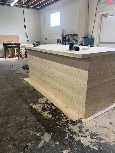 A light natural wood custom made home bar with a thick white top surface, shown in a workshop or studio setting with a concrete floor.