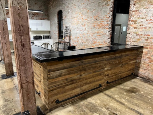 Rustic front view of L shaped reclaimed wood bar with black top and industrial pipe foot rail on concrete floor